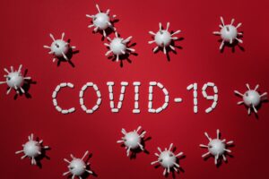 White 3D drawings of coronaviruses are on red background with the words COVID-19 written out in white pill capsules