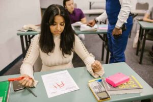 A young woman with dark hair and a cream sweater holds a red marker over her assignment. It looks like she added a line to the paper changing her mark from F+ to A+