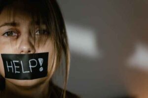 A young woman with blonde hair looks like she is stressed and has black gaffer tape over her mouth with the word "Help!" written on it in white marker.
